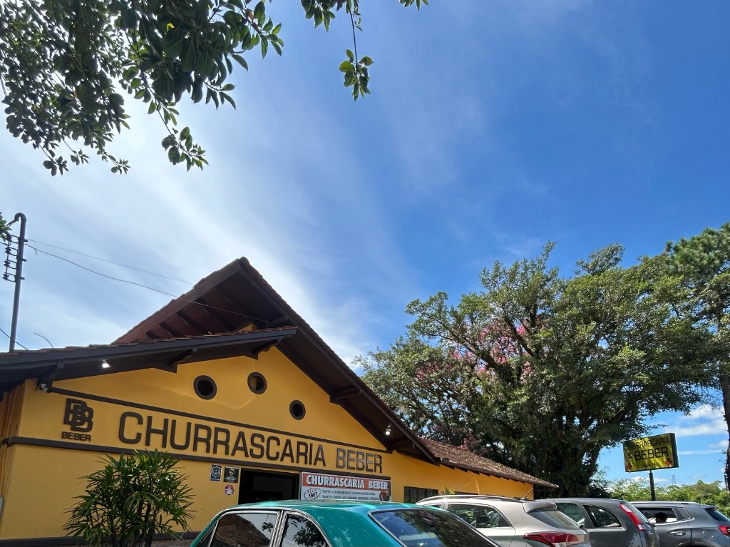 Fachada da Churrascaria Beber com céu, árvores e estacionamento em Ascurra, SC